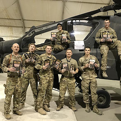 A group of service members holding standing in front of a helicopter in the hanger and holding bags of Williams & Conner jerky.